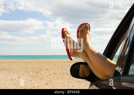 Woman's legs and feet in orange flip flops sticking out of car window in front of empty beach Stock Photo