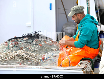 A local fisherman repairs his fishing nets on his boat on quayside in Teulada, South West Sardinia, Italy Stock Photo