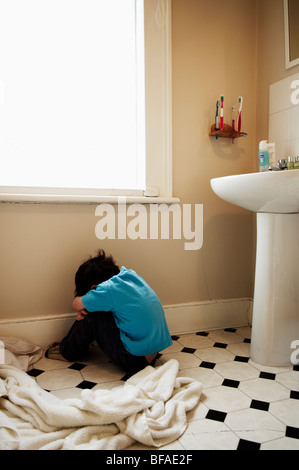 A upset boy sits on the floor of the bathroom with his back turned to camera Stock Photo