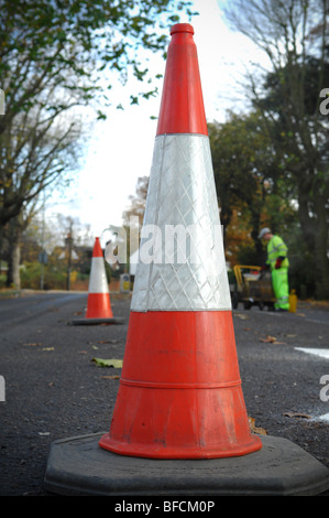 A red and white cone used to direct and contain road traffic in the UK. In the background is a road maintenance worker. Stock Photo