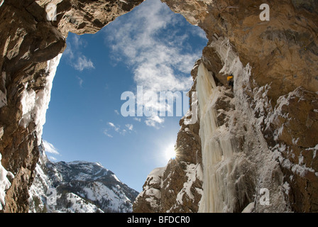 View from below as a professional male ice climber ascends a frozen waterfall on a sheer rock face. Stock Photo
