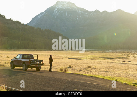 A woman drinks coffee and watches the sunrise from the road next to a field in Yellowstone National Park. Stock Photo