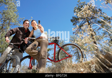 Couple standing with mountain bikes, using mobile phone, photographing selves, low angle view. Stock Photo
