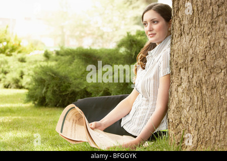 Woman sitting in grass, leaning on tree, holding newspaper. Stock Photo