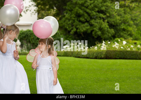 Two young girls in garden, wearing white dresses, holding balloons Stock Photo