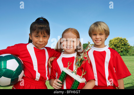 Three children (7-9 years) holding soccer ball and trophy on soccer field, portrait Stock Photo