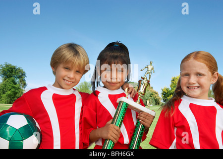 Three children (7-9 years) holding soccer ball and trophy on soccer field, portrait Stock Photo