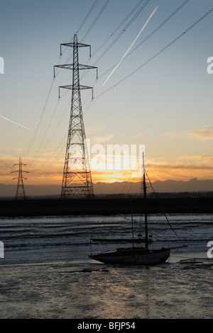 Electricity pylons at sunrise over Faversham creek in Kent. A plane leaves a contrail behind it in the sky. Stock Photo
