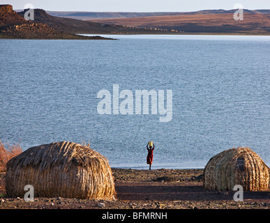 An El Molo woman fetches water from Lake Turkana. Typical dome-shaped El Molo houses in the foreground are made of reeds. Stock Photo