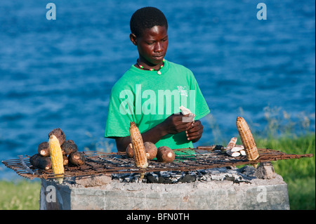 Kenya, Mombasa. A man roasts maize cobs, sweet potatoes and arrowroot on charcoal along the seafront. Stock Photo