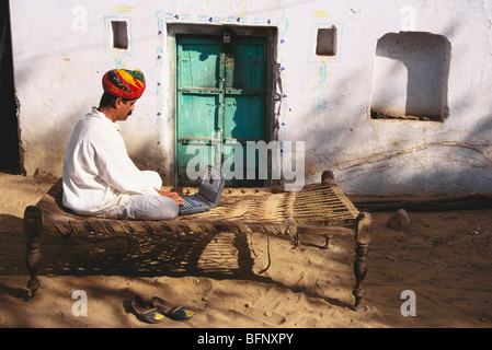 MMN 61890 : Rajput Indian man using laptop computer sitting on khat charpoy outside house ; Pushkar ; Rajasthan ; India Stock Photo