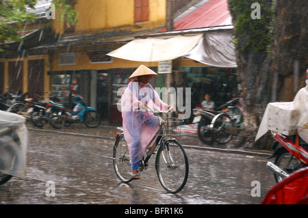 Woman cycling through the pouring rain falling in the streets of Hoi An, a small Vietnamese coastal city Stock Photo