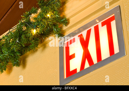 A closeup of an exit sign, lit, with green garland and white Christmas lights beside. Stock Photo