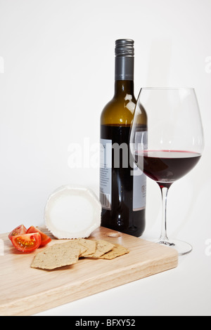 Goats cheese with tomatoes and crackers on a wooden cheese board accompanied by a bottle and glass of red wine. Stock Photo