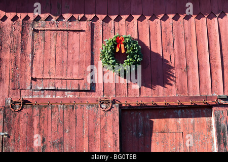 green Christmas wreath with bright red bow hangs above classic barn door on old red wooden barn building in Montgomery New York Stock Photo