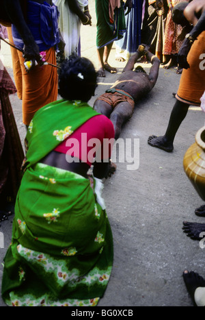 A pilgrim rolls on the ground which is hard pavement completely around the Palani Murugan Temple holding a coconut during the annual Hindu Thaipusam festival. He is assisted by family members who occasionally give her water. This is a form of kavadi which is an act of devotion represented by the hardship and pain she is experiencing. Stock Photo