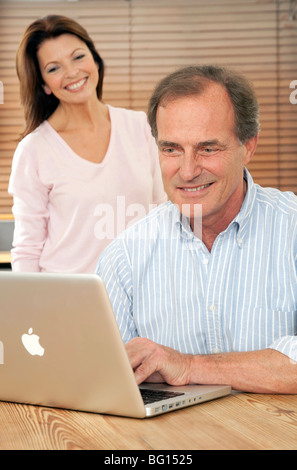 Smiling woman looks at husband or partner as he uses a mac laptop computer at the kitchen table Stock Photo