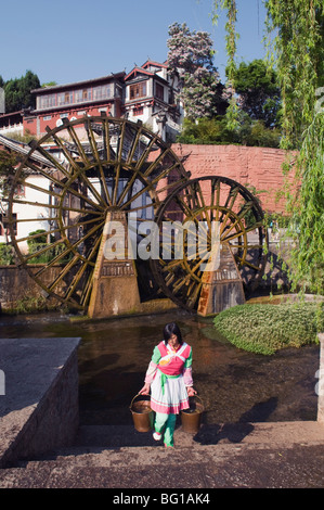A Bai girl carrying buckets of water in front of a water wheel in Lijiang Old Town, UNESCO, Yunnan Province, China, Asia Stock Photo
