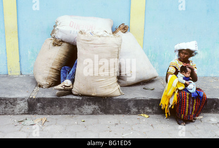 A mother sits on a sidewalk with her sleeping baby while a man sleeps between some large sacks in Joyabaj, Guatemala Stock Photo