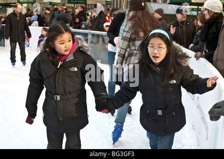 2 tween Asian girls skating on the pond at Bryant Park, one holding tight to the other as she lets go edge of rink support Stock Photo