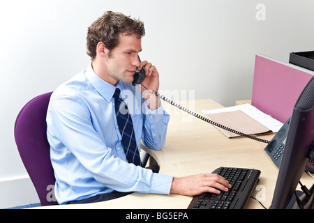 A male office worker answering the phone and looking at a computer monitor in a customer service centre. Stock Photo