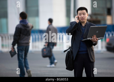 A man makes a call on his mobile phone as he holds an open laptop computer on a roadside in northern Beijing, China. Stock Photo