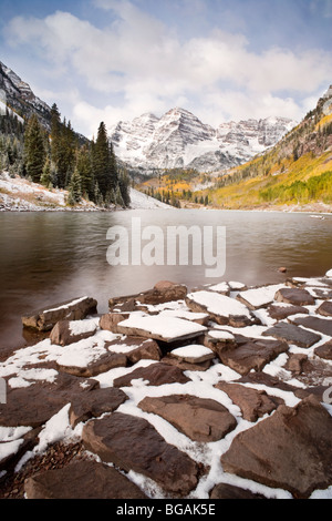 Maroon Bells in the background and large stones with snow along the shores of the lake. Cloudy sky and autumn colours Stock Photo