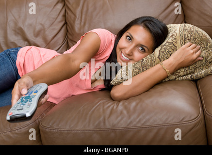 Relaxing on a sofa at home, a young woman plays with the TV remote control. Stock Photo