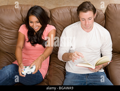 A young couple relax at home in two different ways; one plays a video game and the other reads a book together on the sofa. Stock Photo