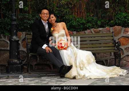 Chinese bride and groom in long wedding gown and dark suit sitting on a bench, Hong Kong, China Stock Photo