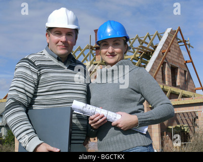Female and male building engineers wearing helmets posing with building plans and laptop against unfinished brick house Stock Photo