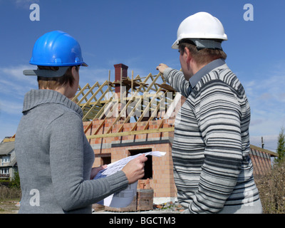 Female and male building engineers wearing helmets discussing building plans over unfinished brick house Stock Photo