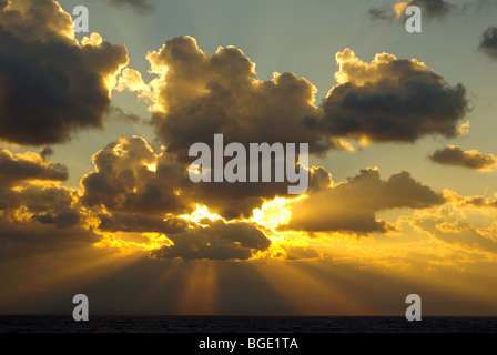 Dramatic sunrise actual (or sunset if required) with sun rays or sunbeam of sunlight bursting through dark clouds and clear blue sky above & beyond Stock Photo