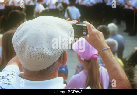 Rear view from above of elderly man wearing flat white cap taking photo on compact digital camera of youth brass band Stock Photo