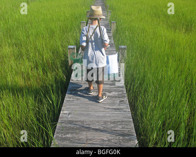 A young boy walking along a wooden pier with fishing pole, net and a bucket. Stock Photo
