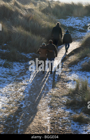 Four people and two dogs walking on a wooden boardwalk through snowy, frosty dunes on New Year's Day 2010 at Burnham Overy. Stock Photo