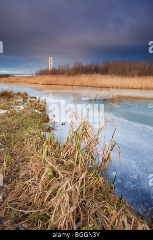 A frozen pond with reeds in the foreground and the Humber Bridge in the distance on a cold winter day Stock Photo