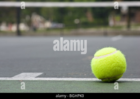 A closeup of a yellow tennis ball just outside of the base line on an asphalt tennis court, with a blurred net in the background Stock Photo