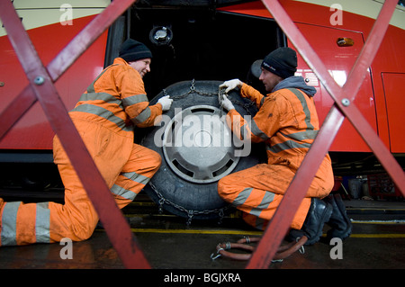 Two mechanics fitting snow chains to a bus wheel using a bus garage inspection pit. Stock Photo