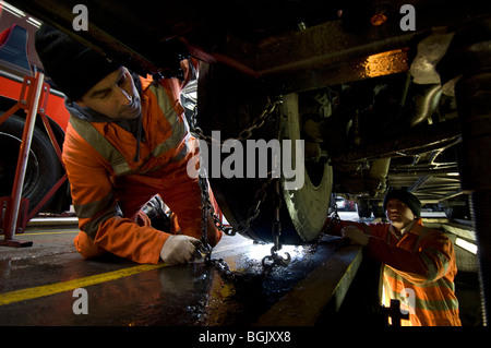 Two mechanics fitting snow chains to a bus wheel using a bus garage inspection pit. Stock Photo