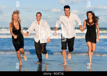 Four young people, two couples, holding hands, having fun and running in the sea on a beach Stock Photo