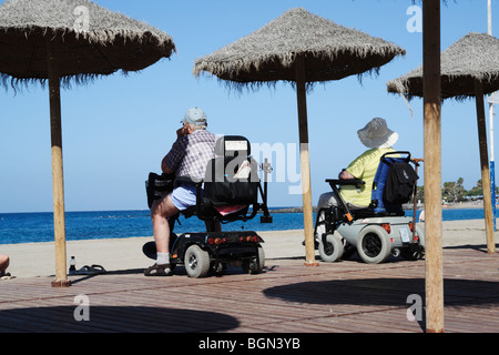 Couple on mobility scooters looking out to sea from Los Cristianos beach on Tenerife in The Canary Islands Stock Photo