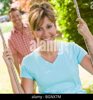 Portrait of a mature woman sitting on a rope swing with a mature man standing in the background Stock Photo