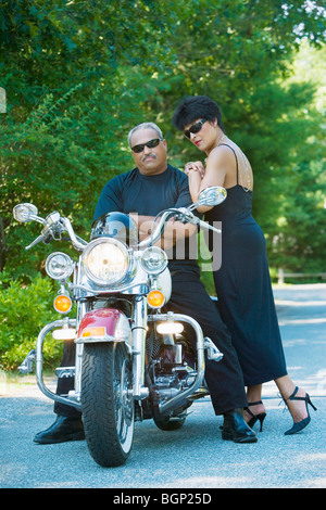 Senior man sitting on a motorcycle with a mature woman standing beside him Stock Photo