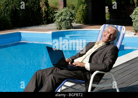 Happy businessman sitting on deck chair with laptop computer next to swimming pool smiling and looking at camera Stock Photo