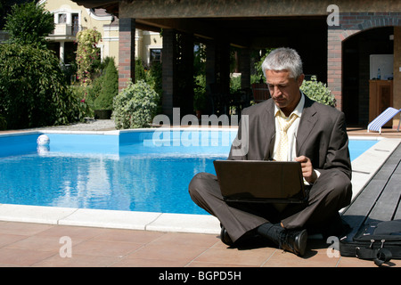 Handsome businessman sitting next to swimming pool with legs crossed using laptop computer Stock Photo