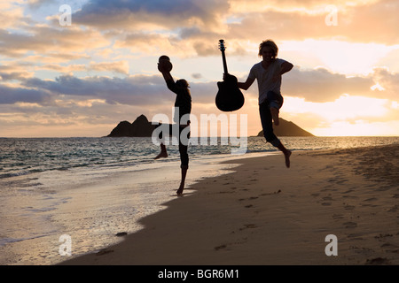 silhouette of twin brothers with drum and guitar leaping in the air at sunrise on a hawaii beach Stock Photo