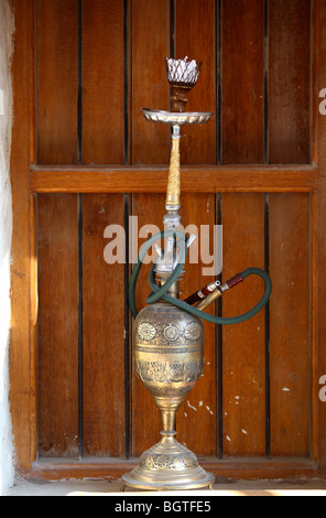 An old hookah or shisha pipe with a brass bowl on a window-cill with a wooden shutter behind, in Souq Waqif, Doha, Qatar Stock Photo