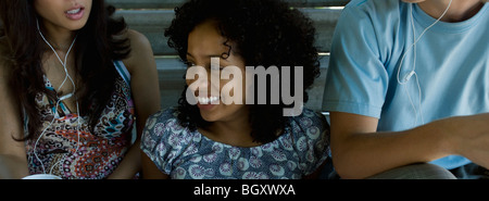 Young woman sitting between two friends listening to earphones, looking away and smiling Stock Photo