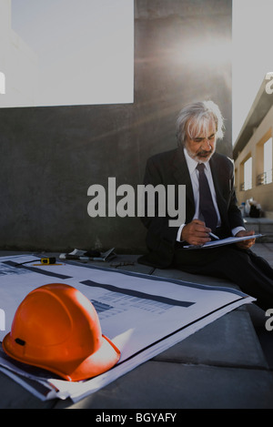 Man sitting on ground at construction site looking at clipboard, hard hat and blueprints in foreground Stock Photo